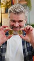 lächelnd kaukasisch Senior Freiberufler Mann Lager Händler halten Abdeckung Augen mit golden Bitcoins BTC. glücklich mittleren Alters männlich Kerl erfolgreich Entwickler Programmierer auf Sofa beim Zuhause Zimmer. zunehmend Reichtum video