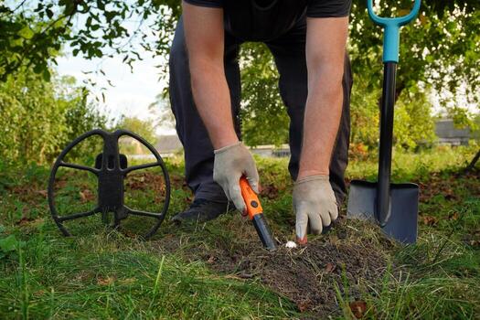 A person searches for treasures in a green garden using a metal detector and a digging tool. The sun sets, casting warm light on the scene, enhancing the tranquil atmosphere photo