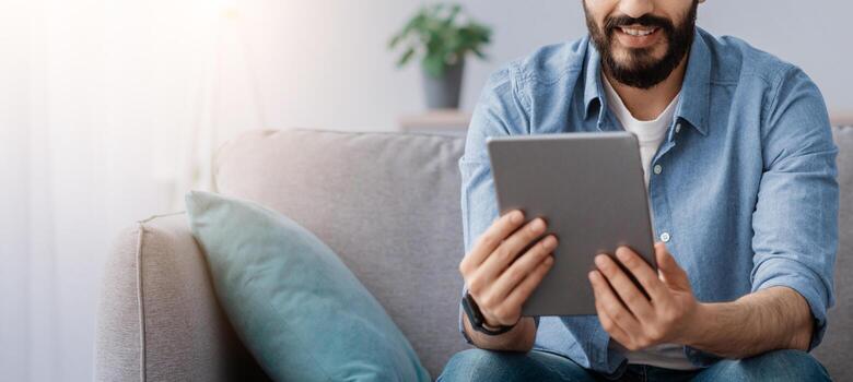 Free time concept. Portrait of happy guy using digital tablet sitting on the sofa at home, free space. Smiling man shopping online, browsing internet, resting on the couch photo