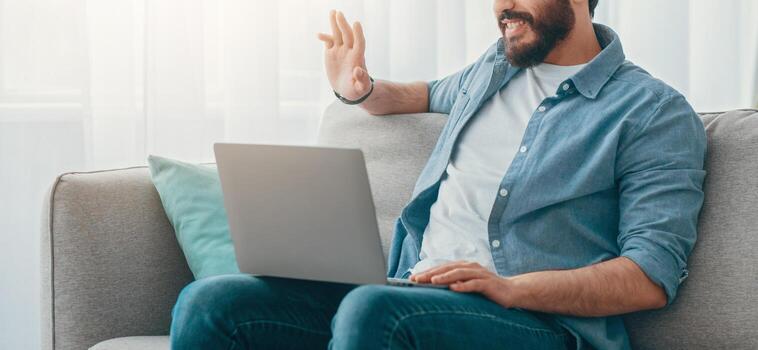 Distant communication. Arab man calling on laptop computer, waving hand to screen while sitting on sofa at home, empty space. Guy communicating with remote friend. photo