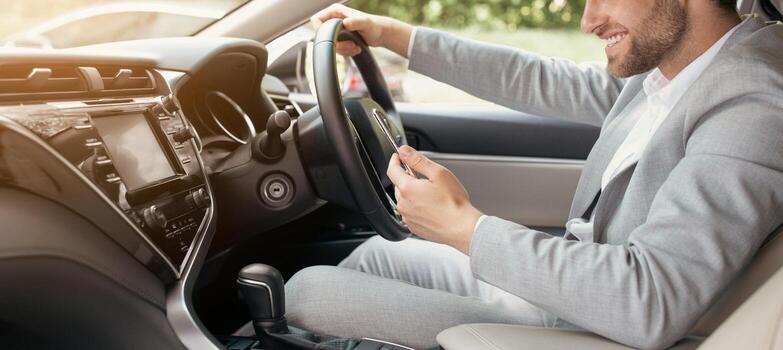Navigation system. Side view of happy handsome businessman using smartphone while sitting in automobile. Rent and trade-in. Young european man in suit is driving in city and looking at gps map photo