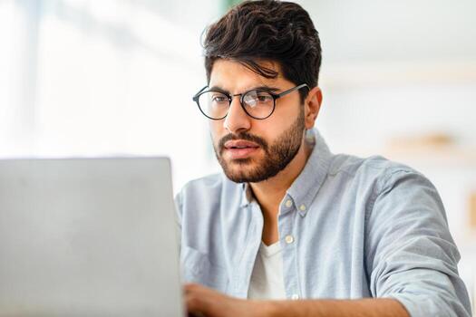 Remote work concept. Concentrated arabic male freelancer working on laptop, typing on computer keyboard, communicating with clients. Man looking at computer screen with pensive face expression photo
