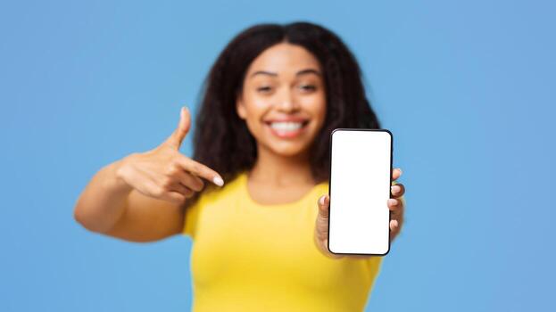 Mockup image of black lady pointing at smartphone with blank screen, demonstrating free place for design or advertisement, recommending app or website, blue studio background photo