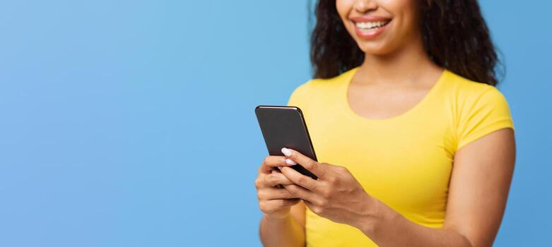 Chatting with friends. Happy black woman using smartphone texting sms message or scrolling social networks feed, standing over blue studio background, selective focus photo