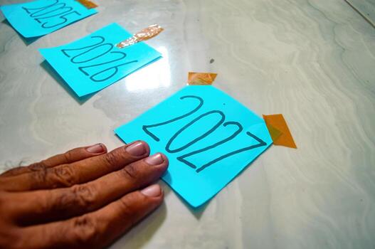 Three blue sticky notes labeled 2025, 2026, and 2027 arranged diagonally on marble surface, with orange tape and hand on 2027 symbolizing future goals and new year focus. photo
