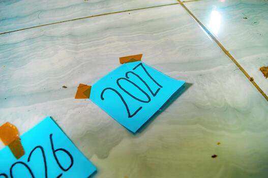 Blue sticky notes labeled 2026 and 2027 taped to tiled floor with brown tape, showing a hand near 2027 symbolizing future goals, planning, and new year focus in a casual setup. photo