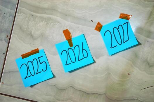 Three blue sticky notes labeled 2025, 2026, and 2027 taped to tile floor with orange tape symbolizing timeline, planning, and new year goals in a creative setup photo