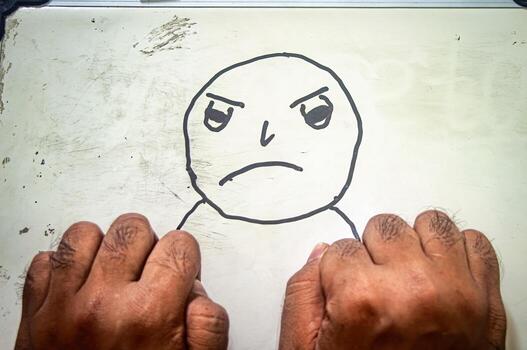 Clenched fists placed below an angry cartoon face on whiteboard blending real gesture with drawn emotion to express frustration, strength, and bold reaction. photo