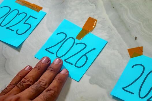 Three blue sticky notes labeled 2025, 2026, and partially 2027 taped to marble surface with orange tape, hand touching 2026 symbolizing timeline, goals, and new year focus. photo