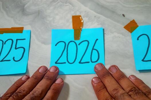 Three blue sticky notes labeled 2025, 2026, and 2027 taped to marble surface with orange tape, hands touching 2026 symbolizing new year focus, timeline, and future planning photo