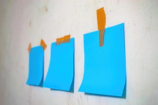 Three blank blue sticky notes taped to a textured white wall with orange tape, ideal for concepts of planning, memory, minimalism, or creative workspace. photo