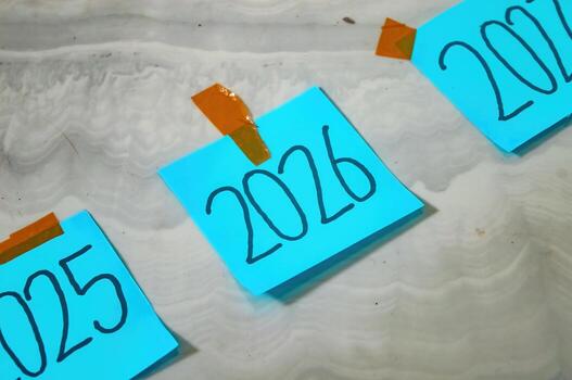 Three blue sticky notes labeled 2025, 2026, and 2027 taped to white marble surface with brown tape symbolizing timeline, planning, and new year goals in a creative layout. photo