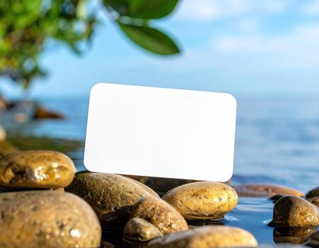Blank white card rests on wet stones by a tranquil lake. Lush foliage and clear sky frame the scene photo