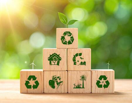 Stacked wooden blocks, each with an eco-friendly symbol, signifying sustainability. A small plant sprouts from the top block. Blurred green background with sunlit bokeh photo