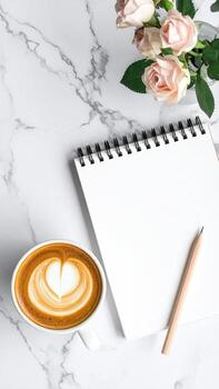 A blank spiral-bound notebook sits beside a latte on a marble surface, with pink roses in the background photo