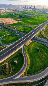 Aerial view of a complex highway interchange, winding roads intersecting over lush green fields, with modern city buildings in the distance photo