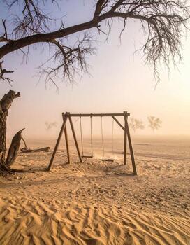 Desolate swing set in a dusty desert landscape. Bare, skeletal trees and a wooden swing set in the middle of a sandy plain. Foggy, light sunrise or sunset. Empty and serene photo