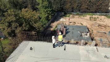 två ingenjörer i hård hattar inspektera konstruktion webbplats från tak, kran krok synlig, över huvudet se av byggnad fundament arbete video