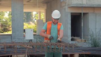 Builder In Helmet And Safety Vest Tying Rebar, Smiling Man Working On Construction Site With Concrete Building Frame In Background, Daytime Work Scene video