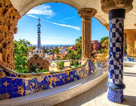 Park bench overlooking city. Mosaic tiled archway with vibrant blue and white patterns framed by pillars. Cityscape with buildings and a tower visible beyond. Sunny day photo