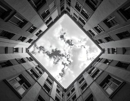 A black and white perspective of a courtyard, looking up at a sky filled with clouds. Buildings surround the central space photo