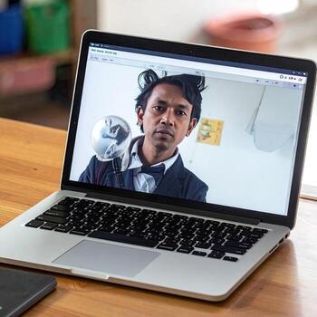 Laptop screen displays a man in a call. Man wears a formal suit and unusual accessory. Light wooden table. Simple background photo