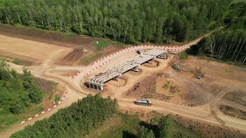 Drone aerial view of a highway bridge under construction in a forest area with earthworks, building materials, and a dump truck driving on a dirt road. video