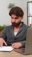 indiano homem às casa Cruz Verificações números em computador portátil e papel notas escreve marcas permanece focado. árabe cara às escrivaninha lê colunas compara totais e valores confirma Combine profundo Reveja calma relaxar humor video
