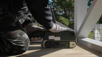 uma trabalhador areias uma de madeira chão em uma terraço usando a elétrico lixadeira dentro brilhante luz solar. video