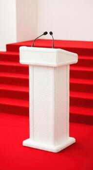 A white podium with microphones stands on a red carpeted stage photo