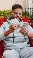 Planung Budget. Reich glücklich indisch Mann verwenden Smartphone Taschenrechner App Anzahl Geld Kasse, Berechnung inländisch Rechnungen Gehalt beim Zuhause Zimmer. Kerl zufrieden von Einkommen Verdienste Sieg spart Geld zum geplant Geschenke video