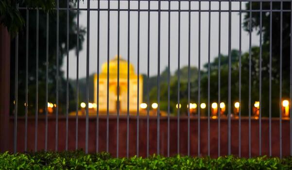 A blurred and colorful monument seen through a close-up of a metallic fence during an evening setting, conveying a sense of focus and perspective with vibrant light. photo