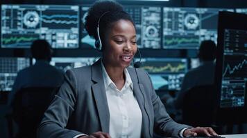 Lycklig skön teknisk kund Stöd specialist har en headsetet ring upp medan arbetssätt på en dator i en mörk övervakning och kontrollera rum fylld med kollegor och visa skärmar video