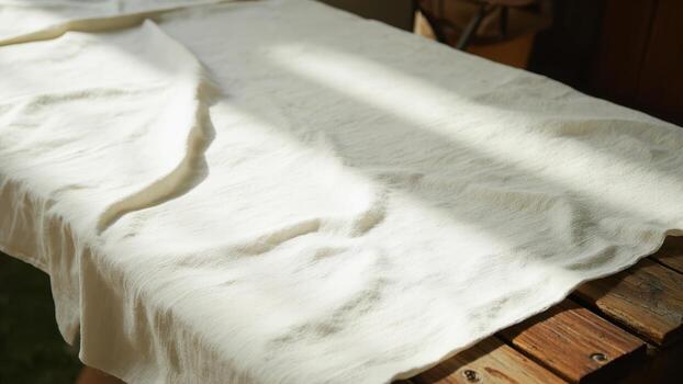 White or beige wrinkled textile draped on a rustic wooden table illuminated by natural sunlight from a window with focus on the fabric texture. Rustic textile and handwoven material concept photo