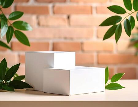 Two blank white boxes are stacked on a light surface with green leaves, against a blurred brick wall background, creating a natural and urban combination. photo