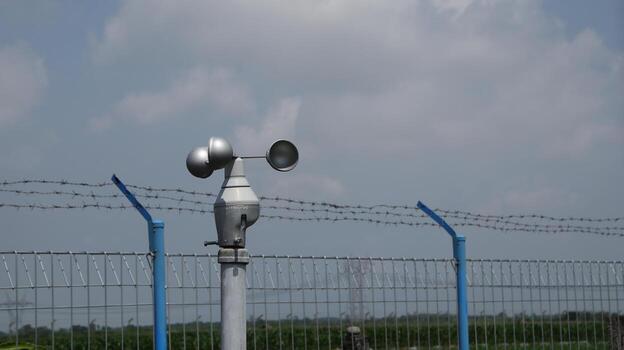 Anemometer measures wind speed near barbed wire fence under cloudy sky, indicating weather monitoring and security measures in a rural area on a bright day. photo