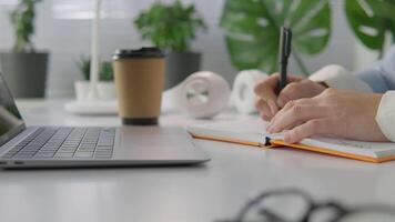 Hände Schreiben im ein Notizbuch beim ein aufgeräumt Arbeitsplatz mit ein Laptop, Kaffee, und Grün, abbilden ein heiter Umgebung zum Fokus und Kreativität video