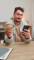 Planung Familie Budget. lächelnd kaukasisch Mann Zählen Geld Euro Kasse verwenden Smartphone Berechnung inländisch Rechnungen. Kerl zufrieden von Einkommen, spart Gehalt Geld beim Zuhause Büro Tabelle Arbeitsplatz. Vertikale video