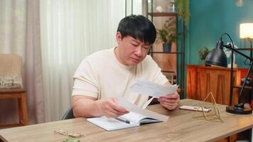 asiatisk man Sammanträde på Hem tabell recensioner verktyg räkningar, rynkar pannan, spottar anteckningar i anteckningsbok, beräknande belopp och tidsfrister. kinesisk kille fokuserar tätt, spårning varje figur för exakt budget. video