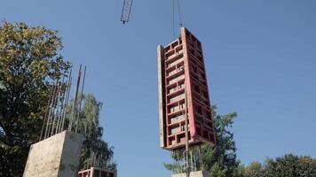 Large red formwork panel lifted by crane at construction site, concrete columns with steel rebars visible against clear blue sky and surrounding trees video