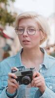 retrato de alegre caucásico hembra viajero en bueno estado animico con cámara en pie en ciudad calle. contento bonito joven rubia mujer turista en lentes sorprendido tomando foto. fotógrafo tomando imágenes video