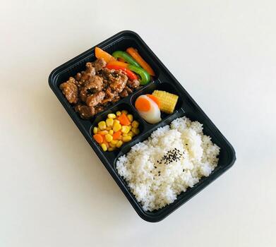 Black bento Box container holds balanced meal of beef stir-fry with vegetables, and white rice in separate compartments. Container is placed on light table background, top view photo