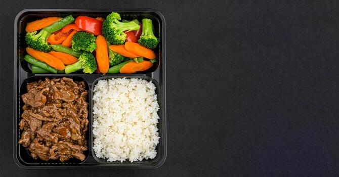 Black bento Box container holds balanced meal of beef stir-fry with vegetables, and white rice in separate compartments. Container is placed on black table background, top view, copy space for text. photo