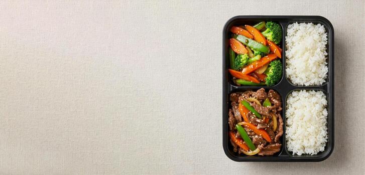 Black bento Box container holds balanced meal of beef stir-fry with vegetables, and white rice in separate compartments. Container is placed on light table background, top view, copy space for text. photo