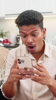 indisch Mann beim Zuhause Uhren Beeindruckend auf Smartphone fühlt sich Schock lacht lächelt aufgeregt durch Nachricht. arabisch Kerl auf Sofa Anteile Verknüpfung im Bote sendet zu freunde schreibt kurz Antworten und Beiträge Reaktion Emoji video