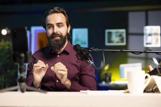 Happy media star using cellphone on tripod to film vlog channel intro for online streaming platforms. Joyful man greets viewers, presenting today topic, recording with mobile phone photo