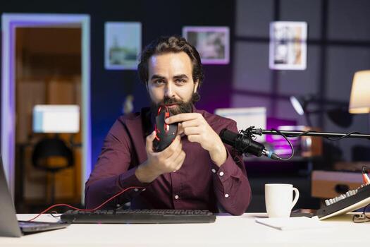 Influencer promoting wired gaming mouse from sponsor to audience in home studio. Content creator paid by company to showcase computer peripheral device, recording with professional microphone photo
