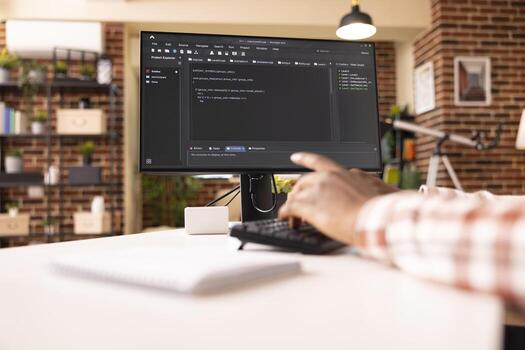 Closeup of computer screen showing programming scripts in progress in brick wall apartment. African American web developer seated at desk, writing code on desktop pc in brick wall home. photo
