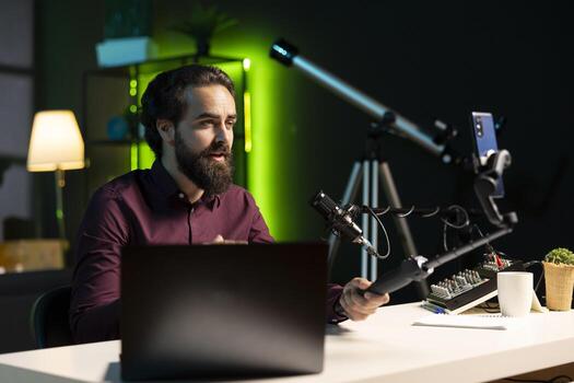 Influencer in home studio recording live broadcast using smartphone on selfie stick, reading messages from fans on laptop. Media star hosting q and a session, filming with phone, answering questions photo