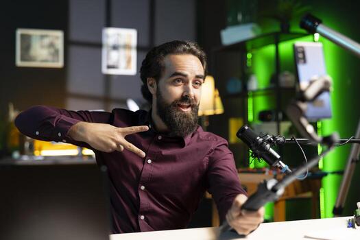 Influencer doing peace sign in front on cellphone attached to selfie stick in studio, chatting with viewers using recording mic. Man doing hand gesturing. using phone and microphone to record vlog photo